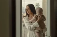 Mulher segura bebê no colo, ambos olham a vista da cidade pela janela.