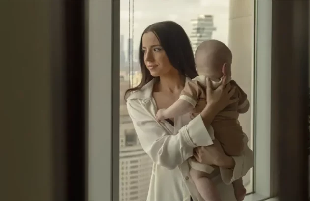 Mulher segura bebê no colo, ambos olham a vista da cidade pela janela.