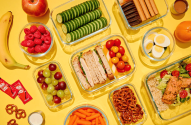 Muitas refeições e lanches pré-preparados em potes de vidro, incluindo sanduíches, saladas e frutas, em fundo amarelo.