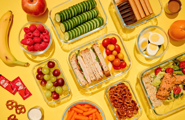 Muitas refeições e lanches pré-preparados em potes de vidro, incluindo sanduíches, saladas e frutas, em fundo amarelo.