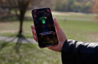 Mão segura smartphone em parque. Tela exibe app com ícone verde de sinal, buscando dispositivo ou conexão.