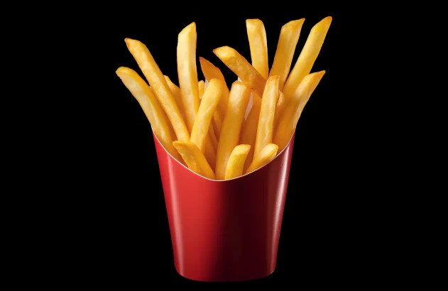 A imagem apresenta uma porção de batatas fritas em um recipiente de papelão vermelho liso, com batatas de tamanho médio e cor dourada, característica do processo de fritura. As batatas estão dispostas de forma a transbordar do recipiente, que apresenta um formato que se expande para cima. O fundo preto destaca as cores das batatas e do papelão. A iluminação uniforme realça o brilho e as texturas das batatas.