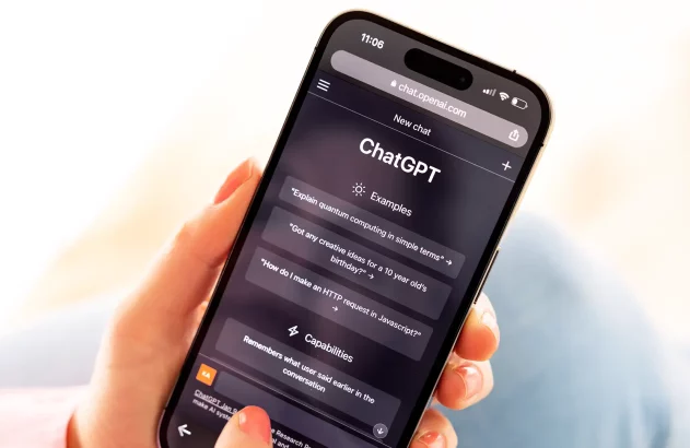 A imagem mostra uma mão com unha pintada de vermelho segurando um celular que exibe a interface do ChatGPT no navegador. Na tela, há seções intituladas "Examples" e "Capabilities," com perguntas e informações sobre o sistema, enquanto o fundo está desfocado.