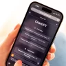 A imagem mostra uma mão com unha pintada de vermelho segurando um celular que exibe a interface do ChatGPT no navegador. Na tela, há seções intituladas "Examples" e "Capabilities," com perguntas e informações sobre o sistema, enquanto o fundo está desfocado.