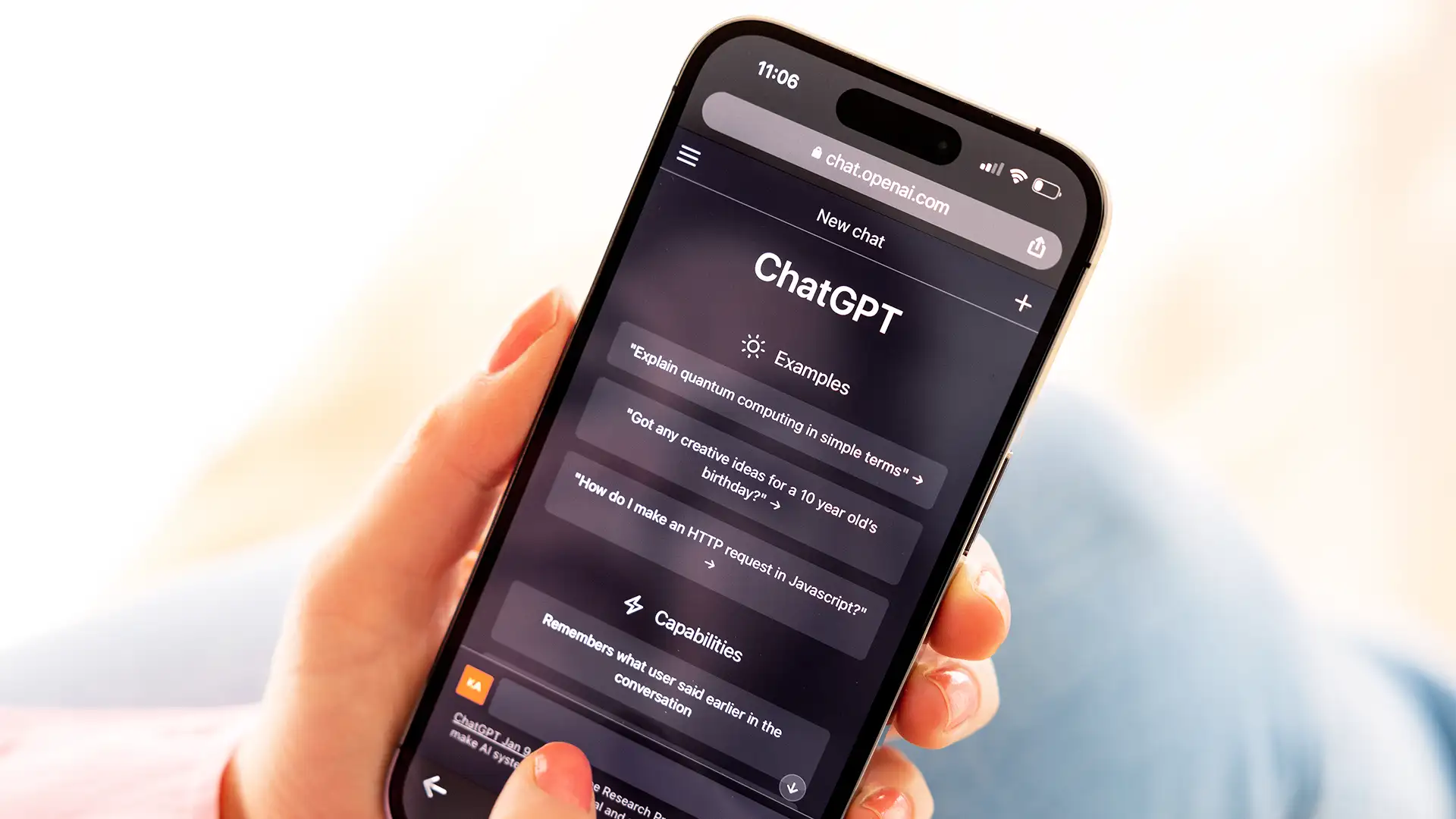 A imagem mostra uma mão com unha pintada de vermelho segurando um celular que exibe a interface do ChatGPT no navegador. Na tela, há seções intituladas "Examples" e "Capabilities," com perguntas e informações sobre o sistema, enquanto o fundo está desfocado.
