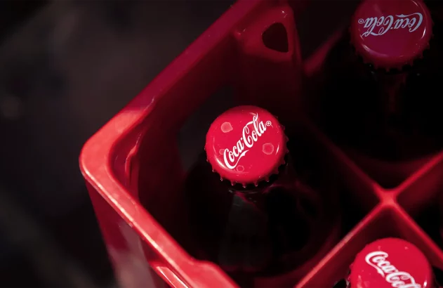 Garrafas de vidro de Coca-Cola com tampas vermelhas posicionadas em um engradado plástico vermelho, visualizadas de cima. A marca Coca-Cola é visível nas tampas das garrafas.