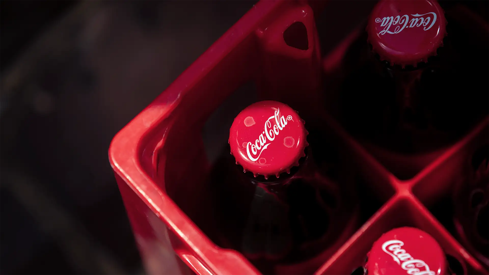 Garrafas de vidro de Coca-Cola com tampas vermelhas posicionadas em um engradado plástico vermelho, visualizadas de cima. A marca Coca-Cola é visível nas tampas das garrafas.