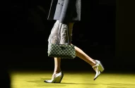 A imagem destaca uma mulher em passarela vestindo um casaco cinza, saia metálica, bolsa geométrica verde/preta e sapatos brancos.