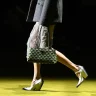 A imagem destaca uma mulher em passarela vestindo um casaco cinza, saia metálica, bolsa geométrica verde/preta e sapatos brancos.
