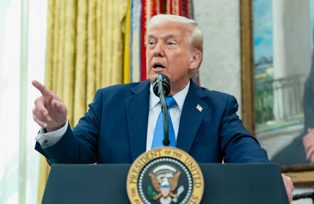 Donald Trump, de terno azul, fala em pódio presidencial e aponta.
