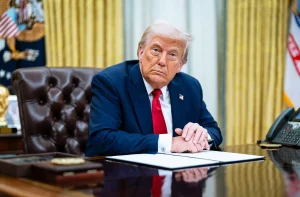 Donald Trump, de terno e gravata vermelha, sentado à mesa no Salão Oval com papéis e telefone.