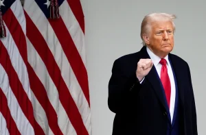 Donald Trump, punho cerrado, de casaco escuro e gravata vermelha, com bandeiras americanas ao lado.