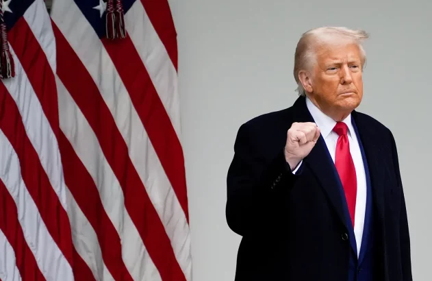Donald Trump, punho cerrado, de casaco escuro e gravata vermelha, com bandeiras americanas ao lado.