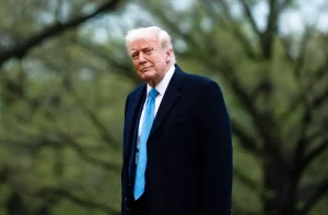 Donald Trump, de casaco azul e gravata azul-clara, em pé ao ar livre com folhagem verde desfocada.