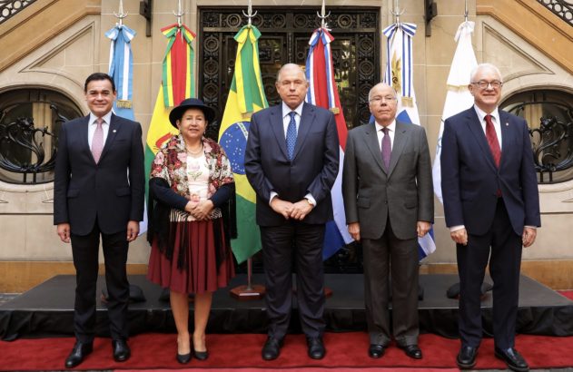 Cinco pessoas (quatro homens, uma mulher de traje típico) posam diante de bandeiras sul-americanas.