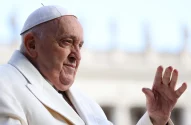 Papa Francisco, de vestes brancas e solidéu, sorri e acena com a mão direita levantada.
