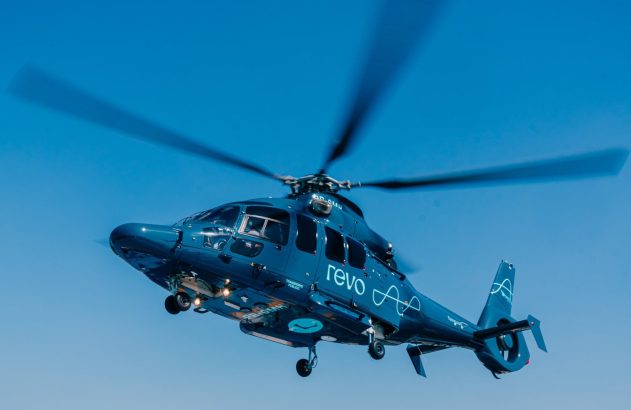 Helicóptero azul da Revo em voo, com hélices borradas, em um céu azul.