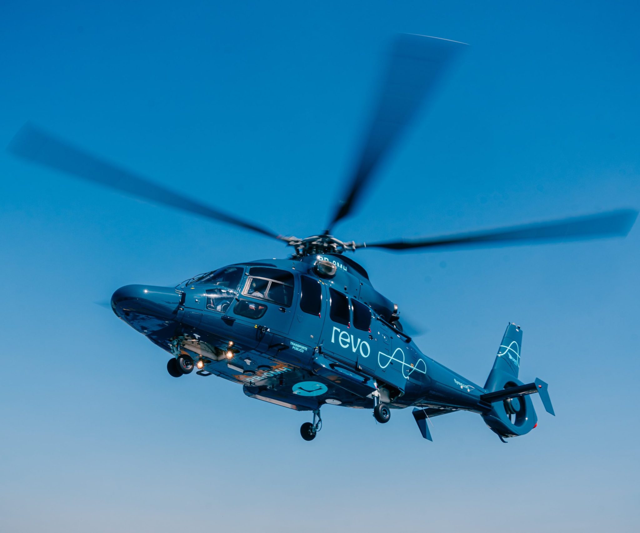 Helicóptero azul da Revo em voo, com hélices borradas, em um céu azul.