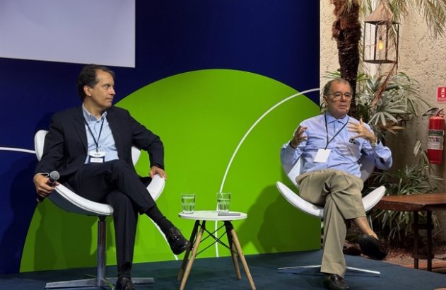 Dois homens em um painel de discussão. Um segura microfone, o outro gesticula enquanto fala. Fundo azul e verde.
