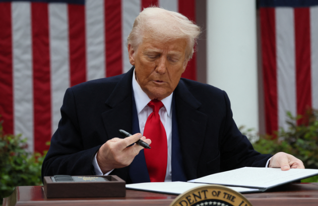 Donald Trump assina um documento em uma mesa, com bandeiras dos EUA ao fundo.