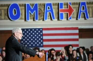 Warren Buffett discursa em pódio. Ao fundo, bandeira dos EUA e o letreiro "OMAHA" com uma seta no lugar do H.