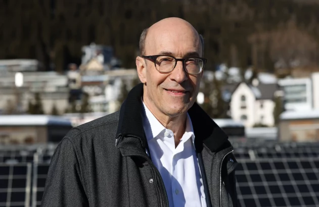 Homem careca com óculos e jaqueta sorri para a câmera. Ao fundo: painéis solares e montanhas nevadas.