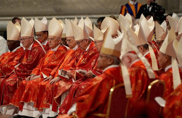 Cardeais ou bispos vestidos de vermelho e mitra branca, sentados em fileiras durante cerimônia.