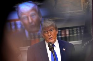 Donald Trump fala ao microfone. Ao fundo, uma projeção borrada de seu rosto e estantes de livros.