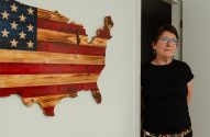 Mulher de óculos e camisa preta, olhando para o lado, emerge de um batente. À esquerda, bandeira dos EUA de madeira em formato de mapa.
