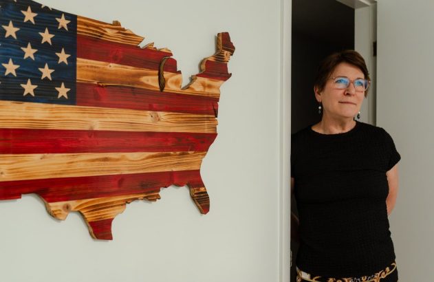 Mulher de óculos e camisa preta, olhando para o lado, emerge de um batente. À esquerda, bandeira dos EUA de madeira em formato de mapa.