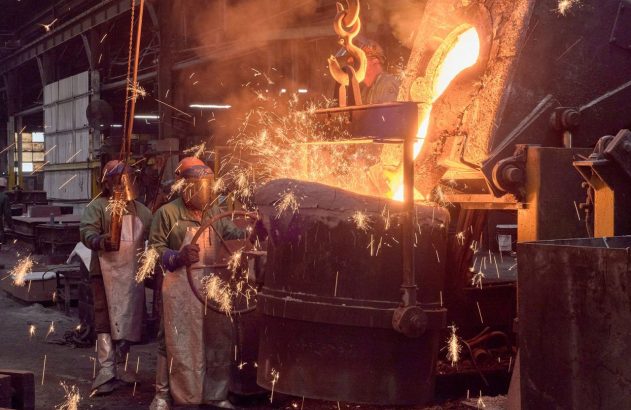 Fundição: Metal incandescente é despejado de uma concha industrial, gerando faíscas e fumaça, com trabalhadores operando.