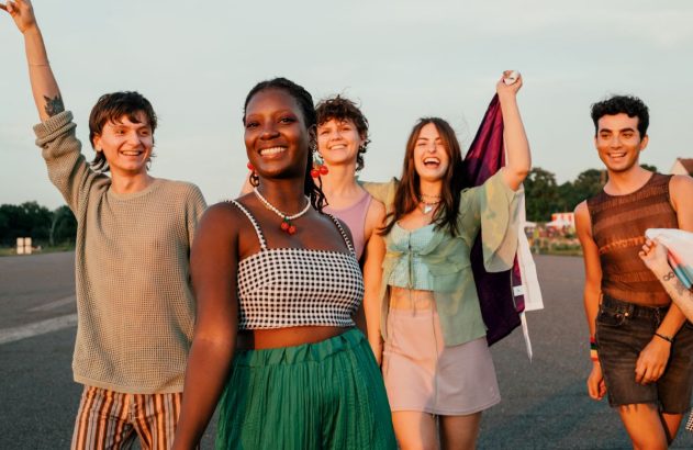Jovens diversos e sorridentes celebram ao ar livre, com braço levantado e bandeira LGBTQ+.