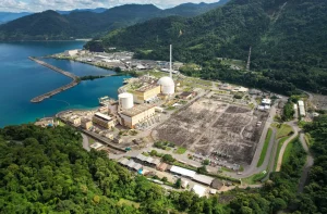 Usina nuclear em complexo industrial costeiro, com reatores, subestação e píer, cercada por floresta e montanhas.