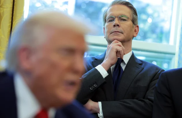 Homem de terno e óculos, mão no queixo, observa Donald Trump desfocado em primeiro plano, que fala.
