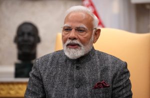 Narendra Modi, com barba branca e óculos, em jaqueta escura, falando em ambiente interno.