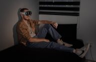 Homem ruivo sentado em um quarto escuro, usando óculos de realidade virtual. A luz lateral ilumina o rosto e o aparelho.
