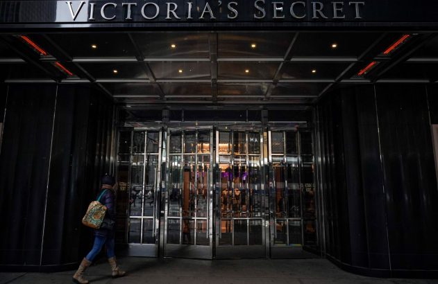Mulher caminha diante da entrada de vidro da loja Victoria's Secret, com o nome da marca visível acima.