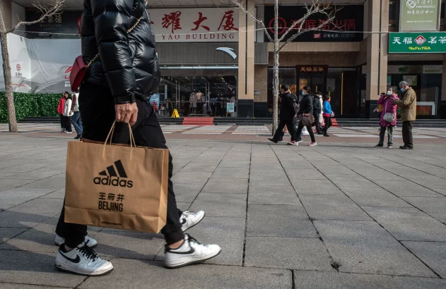 Pessoa em rua de Pequim com sacola Adidas. Outras pessoas transitam ao fundo, algumas de máscara.