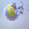 Imagem de balão metálico prateado com símbolo de Bitcoin amarelo no centro, prestes a explodir. Fragmentos são projetados para fora contra um fundo azul-claro.