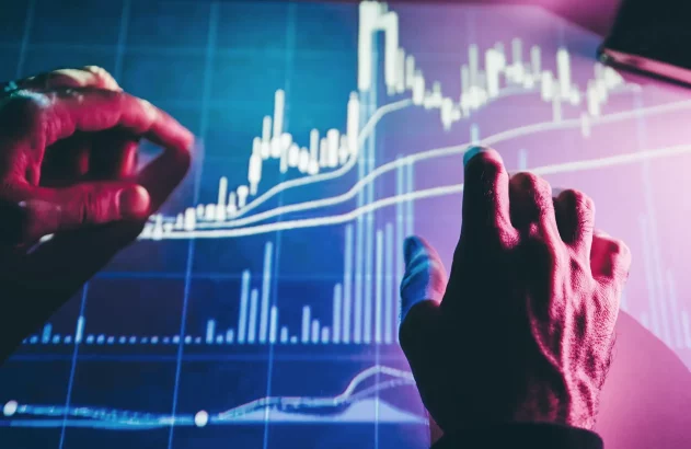 Imagem de duas mãos interagindo com uma tela eletrônica exibindo gráfico financeiro com linhas e barras nas cores branca e azul.