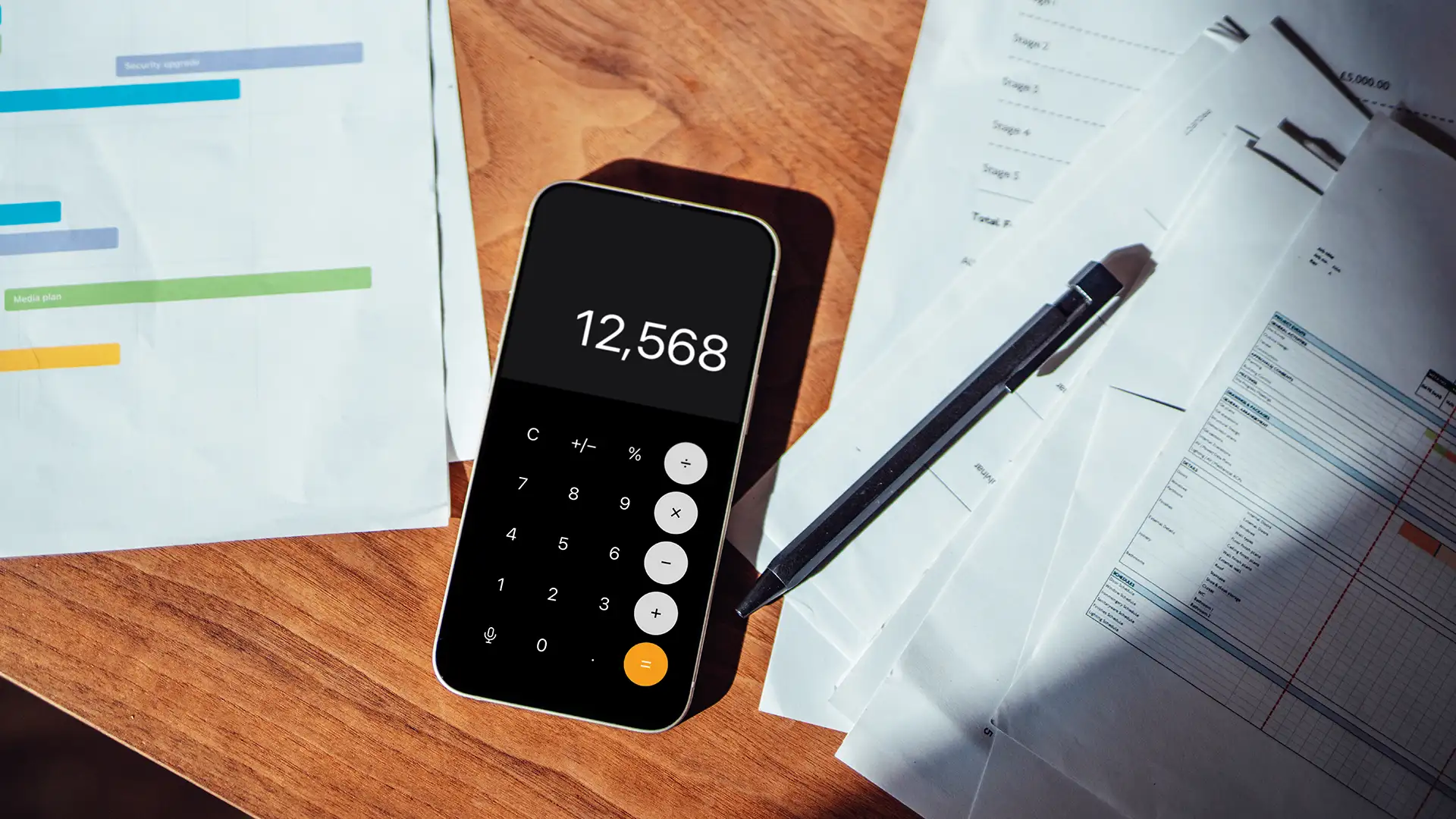 Ambiente de escritório com mesa de madeira, documentos e smartphone. Smartphone mostra tela de calculadora com número 12.568, cercado por papéis, gráficos coloridos e caneta preta.