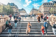 A imagem apresenta uma cena de Paris, mostrando pessoas subindo e descendo uma ampla escadaria. Ao fundo, há edifícios de arquitetura clássica em tons de bege e cinza, e uma rua cheia de veículos em movimento.