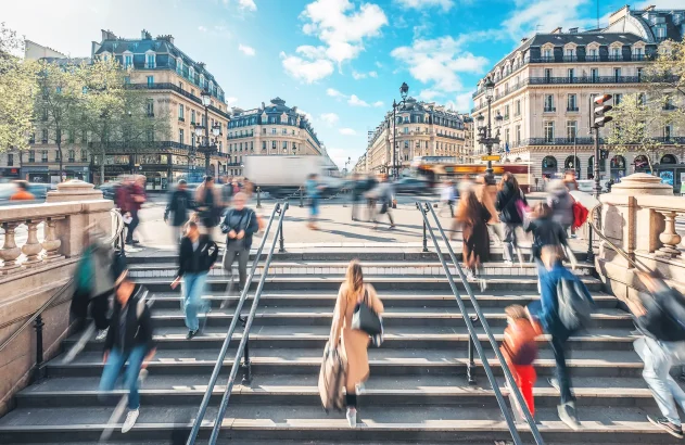 A imagem apresenta uma cena de Paris, mostrando pessoas subindo e descendo uma ampla escadaria. Ao fundo, há edifícios de arquitetura clássica em tons de bege e cinza, e uma rua cheia de veículos em movimento.