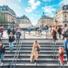 A imagem apresenta uma cena de Paris, mostrando pessoas subindo e descendo uma ampla escadaria. Ao fundo, há edifícios de arquitetura clássica em tons de bege e cinza, e uma rua cheia de veículos em movimento.