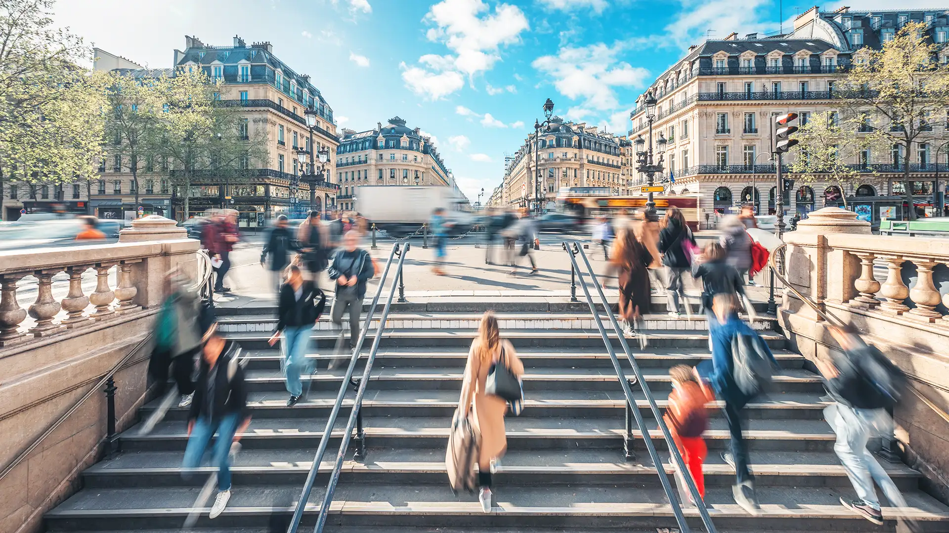 A imagem apresenta uma cena de Paris, mostrando pessoas subindo e descendo uma ampla escadaria. Ao fundo, há edifícios de arquitetura clássica em tons de bege e cinza, e uma rua cheia de veículos em movimento.
