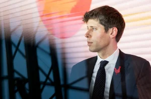 Sam Altman, CEO da OpenAI, de perfil com terno escuro, camisa branca e gravata preta com bolinhas brancas, broche de galo vermelho. Fundo listrado em laranja, amarelo e branco, com círculo laranja, e silhueta de estrutura metálica à frente.