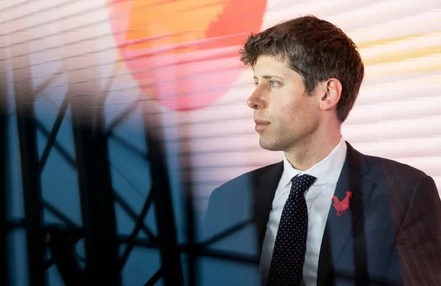 Sam Altman, CEO da OpenAI, de perfil com terno escuro, camisa branca e gravata preta com bolinhas brancas, broche de galo vermelho. Fundo listrado em laranja, amarelo e branco, com círculo laranja, e silhueta de estrutura metálica à frente.