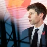 Sam Altman, CEO da OpenAI, de perfil com terno escuro, camisa branca e gravata preta com bolinhas brancas, broche de galo vermelho. Fundo listrado em laranja, amarelo e branco, com círculo laranja, e silhueta de estrutura metálica à frente.