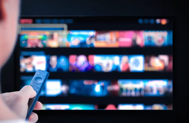 Imagem de uma pessoa com controle remoto apontando para televisão desfocada. Tela exibe miniaturas coloridas organizadas, semelhante a serviço de streaming.