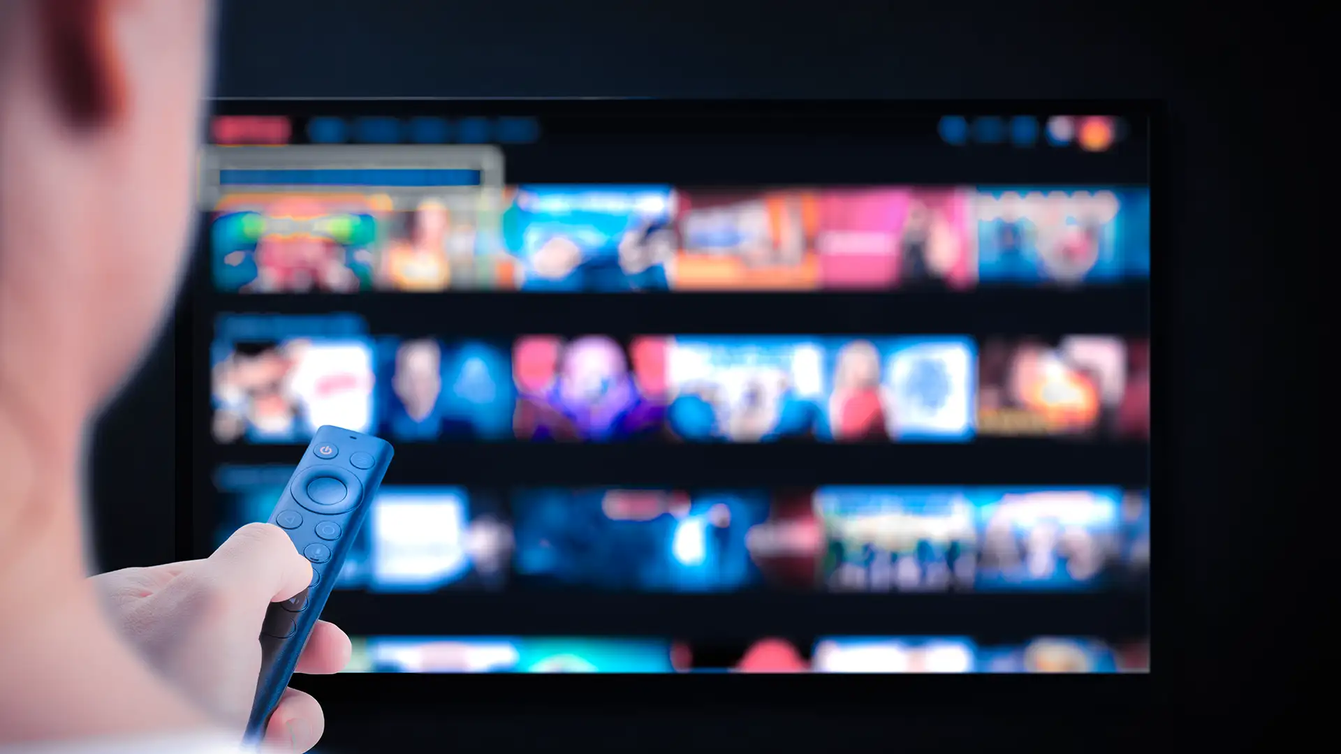 Imagem de uma pessoa com controle remoto apontando para televisão desfocada. Tela exibe miniaturas coloridas organizadas, semelhante a serviço de streaming.
